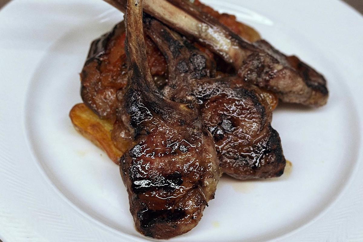 Grilled lamb chops with visible grill marks are stacked on a white plate, served over golden roasted potato wedges.