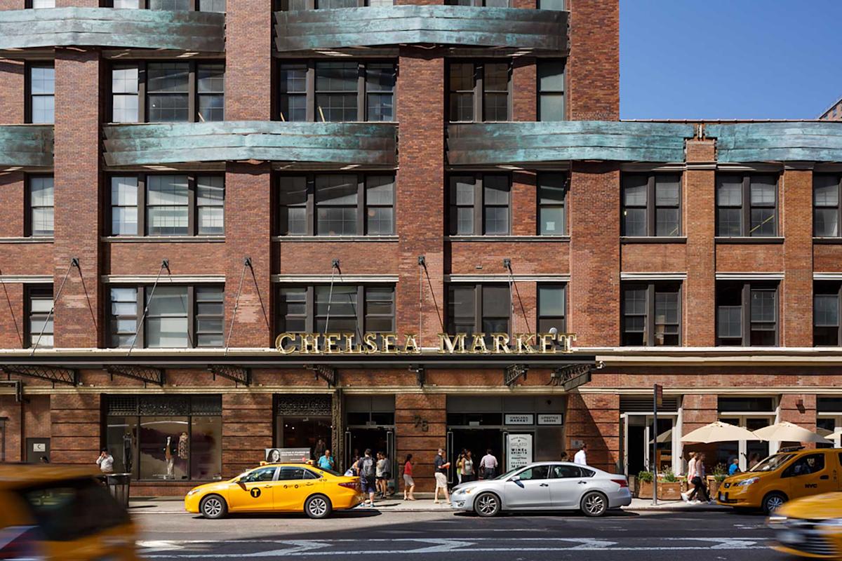Street view of the Chelsea Market building with large windows and a sign above the entrance. Yellow taxis and other cars are passing by, and people are near the entrance and outdoor seating area.