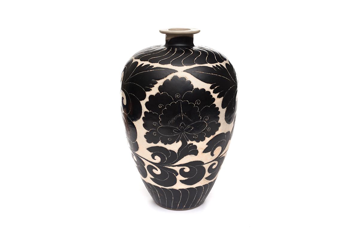 A tall ceramic vase with a narrow neck, featuring bold black floral and leaf patterns on a white background. The design is symmetrical and ornate, and the vase stands on a plain surface with a white backdrop.