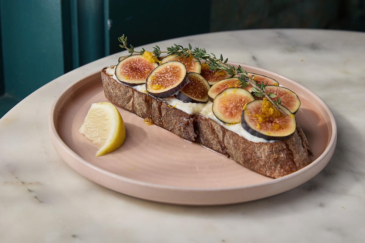 A slice of rustic bread topped with fresh figs, creamy spread, honey, and herbs sits on a pink plate with a lemon wedge on the side, placed on a marble table.