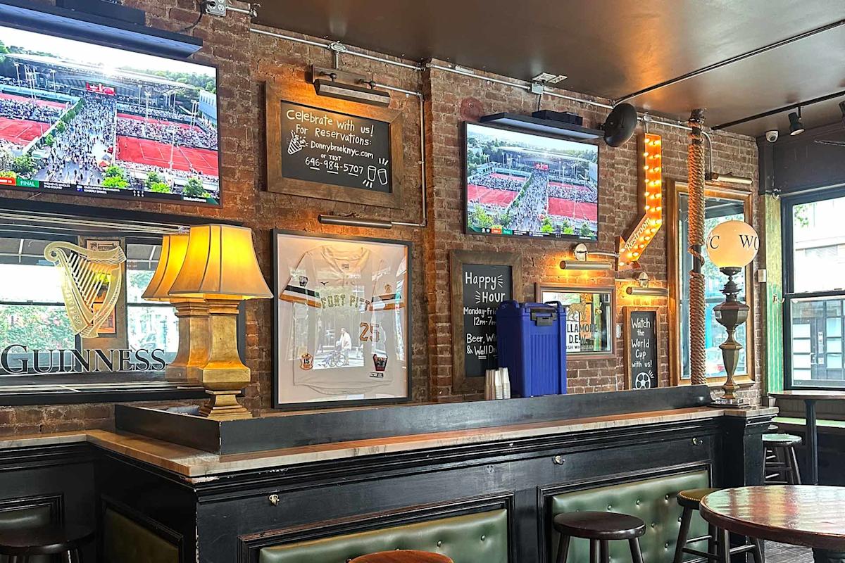 A cozy bar with exposed brick walls, three TVs showing a tennis match, framed sports memorabilia, Guinness signs, lamps, a neon "BAR" sign, and green cushioned seating. Daylight streams through a window.