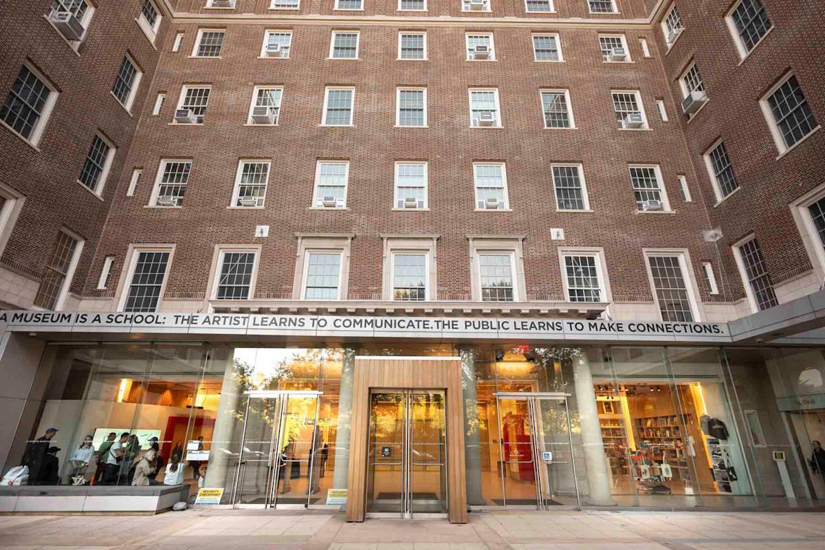 Exterior view of a three-story brick building with large windows. The entrance has glass doors and a quote above them reads, "A museum is a school: The artist learns to communicate. The public learns to make connections.