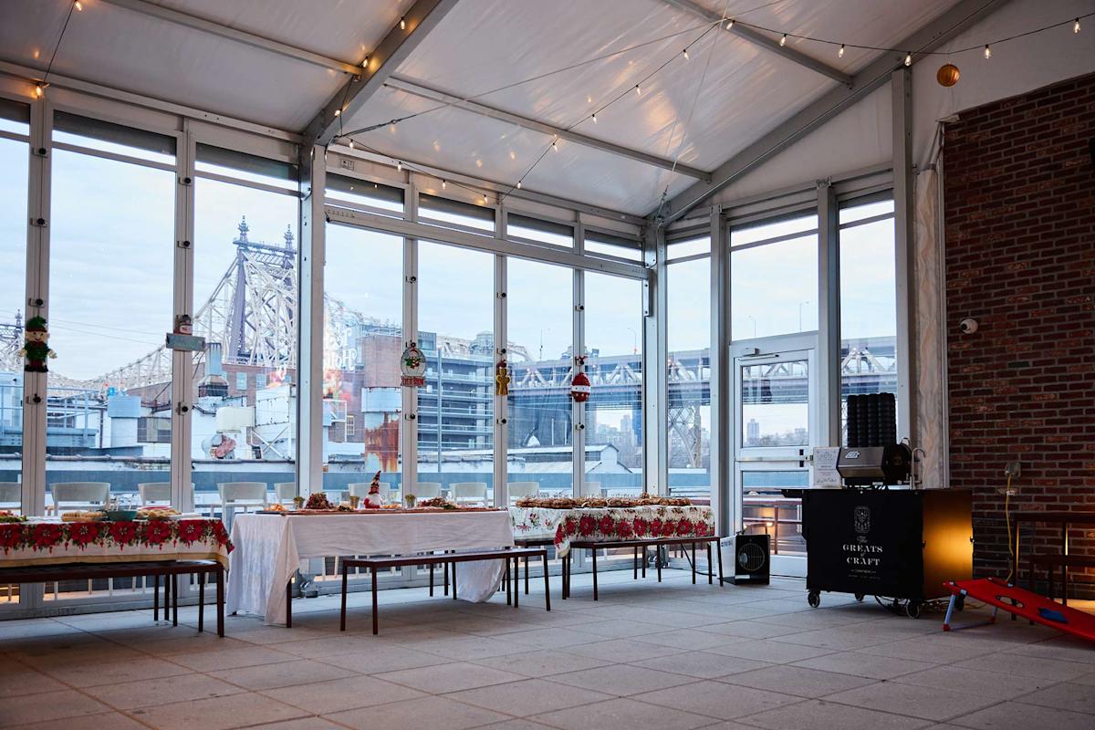 A decorated indoor terrace with string lights, large windows showing a city bridge, and a long table set for a festive event with holiday decorations and food trays.
