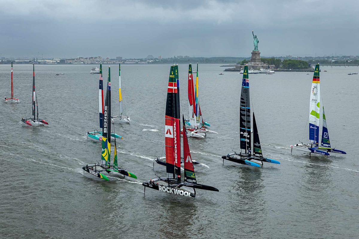Several colorful sailboats race on a gray, cloudy day in a wide harbor, with the Statue of Liberty visible in the background on the right side.