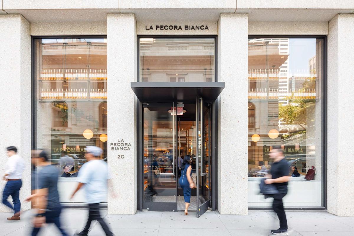 People walk past the entrance of La Pecora Bianca restaurant, which features large windows and a modern facade. One person is entering through the glass door, and the interior is visible with diners inside.