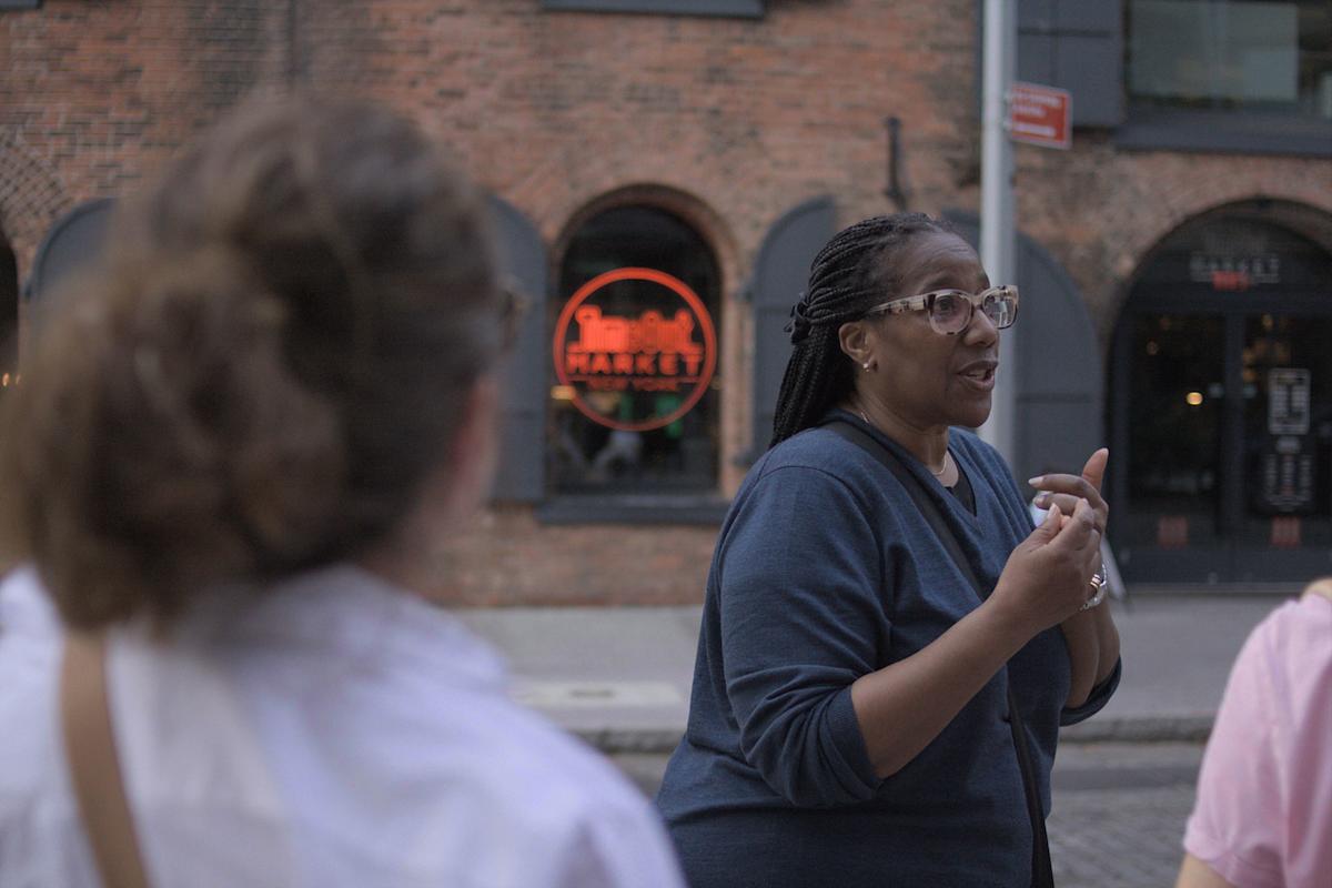 A woman with glasses and braided hair speaks to a small group outside near a brick building with a neon market sign in the window. One person is partially visible in the foreground, listening.