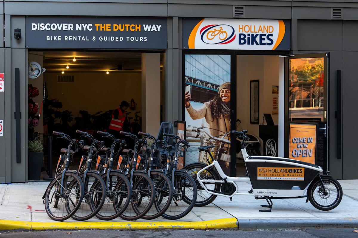 A row of rental bikes and a cargo bike are parked outside Holland Bikes, a bike rental and tour shop in NYC. The storefront features large signs, and a woman is pictured riding a bike on a poster by the entrance.