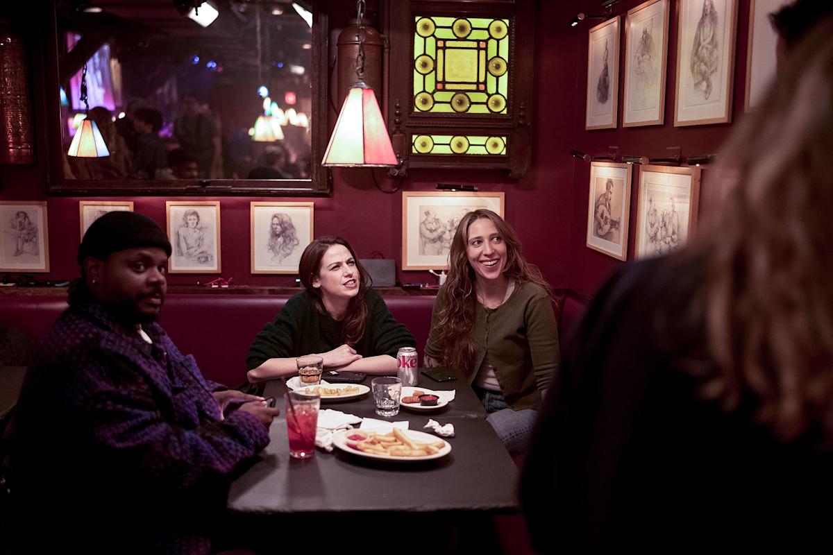 Three people sit at a table in a dimly lit restaurant with framed sketches on red walls, eating and drinking. Two women smile at someone standing, while a man looks toward the camera.