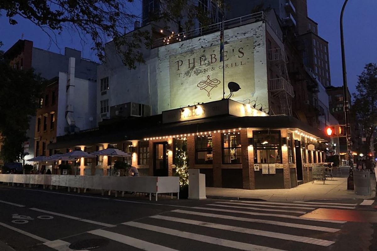 A warmly lit restaurant called Phebe’s Tavern & Grill sits on a street corner at dusk, with string lights, outdoor seating, and surrounding buildings under a darkening sky.