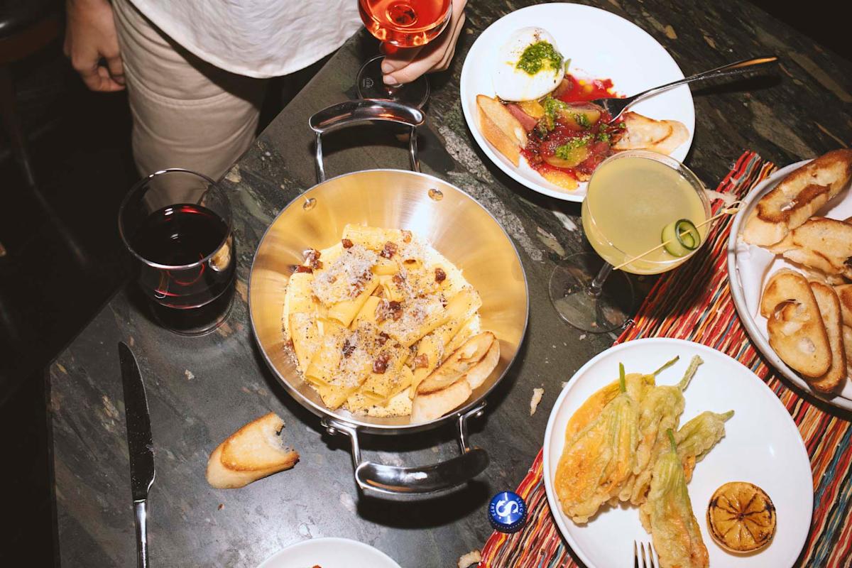 A table set with pasta in a pan, drinks, bread slices, fried zucchini flowers, a burrata salad, and a person holding a glass of rosé. The scene is lively and casual, with food and drinks ready to enjoy.