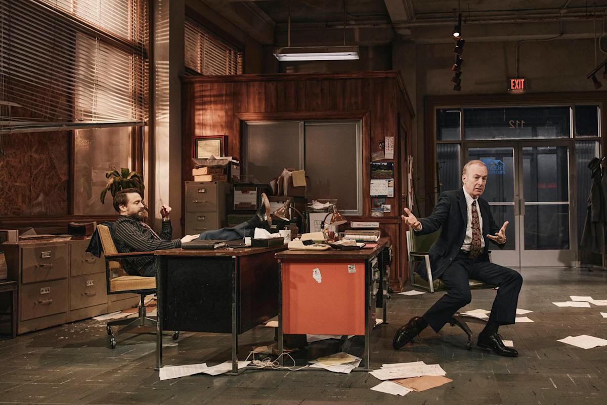 Two men in a messy office: one sits casually with feet on the desk, while the other gestures animatedly in a suit, leaning back in his chair. Papers and files are scattered on the floor and desks.