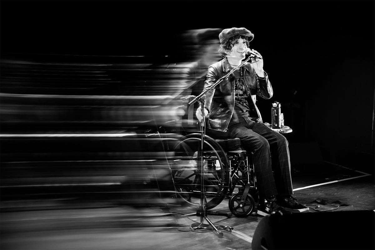 A person in a wheelchair performs on stage, holding a microphone and wearing a hat and leather jacket. Motion blur to the left suggests dynamic movement or energy contrasting with the seated performer. Black and white photo.