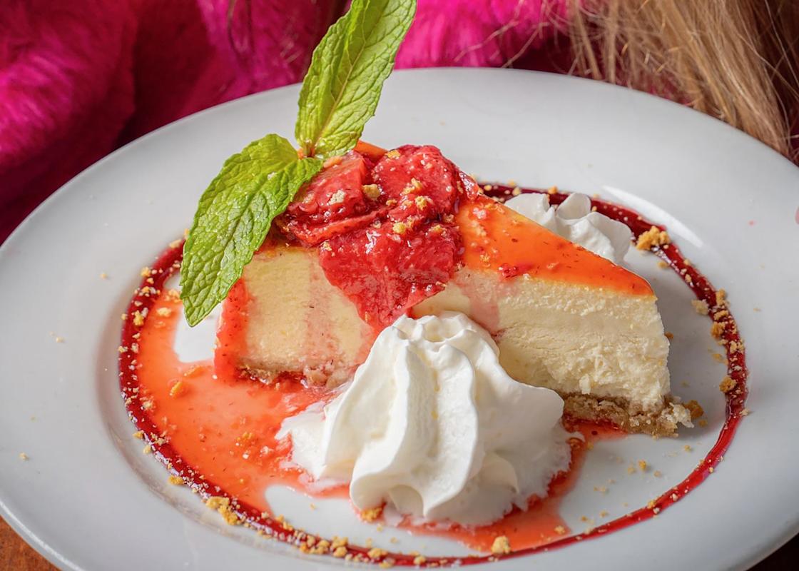 A slice of cheesecake topped with strawberry sauce and a mint leaf, served with whipped cream on a white plate. The plate has sauce drizzled around the dessert.