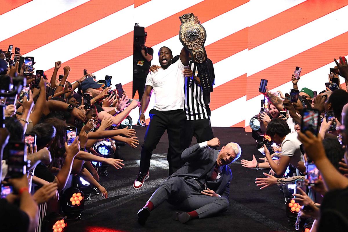 A cheering crowd lines a walkway as a man in a referee shirt holds up a championship belt. Another man celebrates while a third person in a suit lies on the ground, surrounded by fans taking photos.