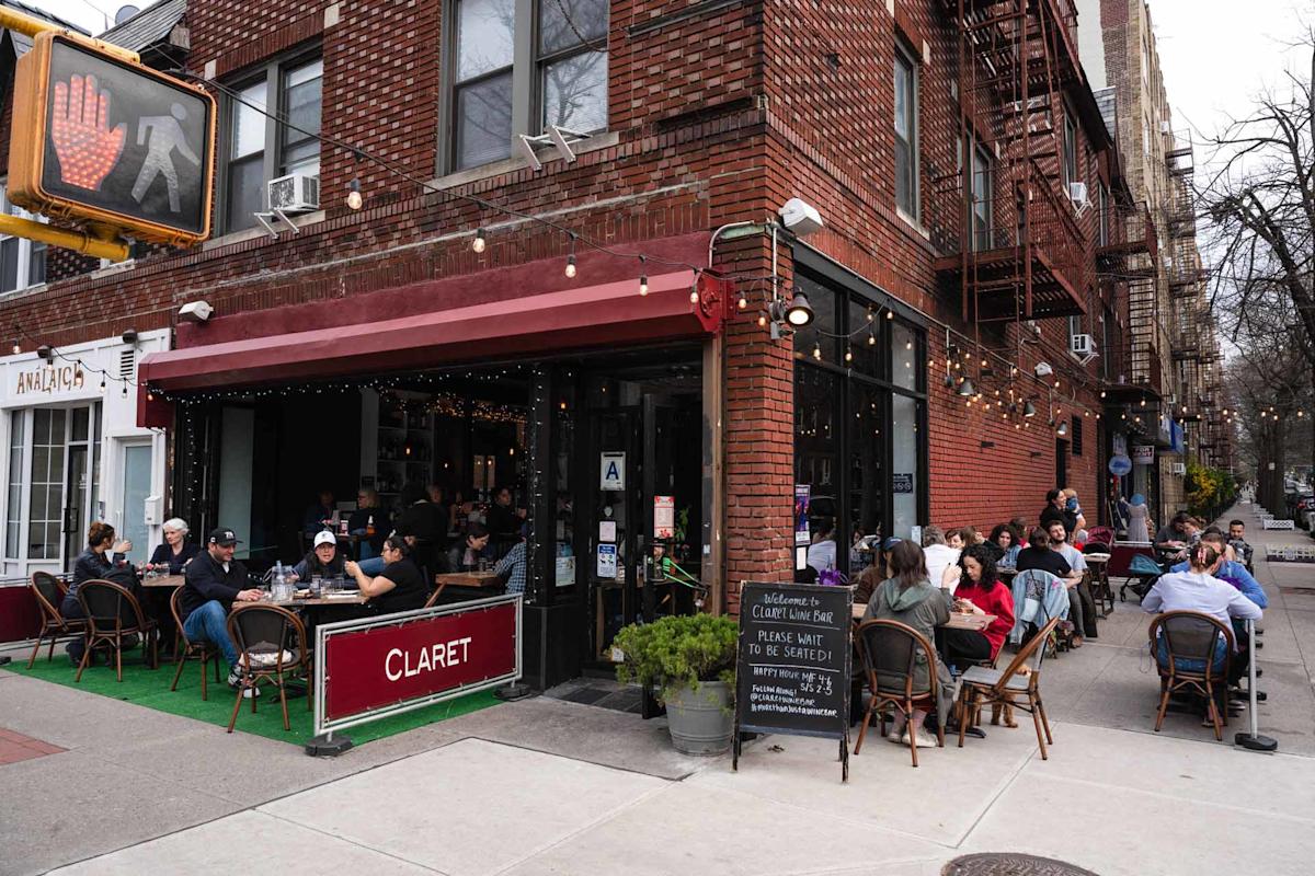 People dine outdoors at a sidewalk café called Claret on a city corner. The café has string lights, a red awning, and a chalkboard menu. A traffic signal and brick apartment buildings are visible in the background.