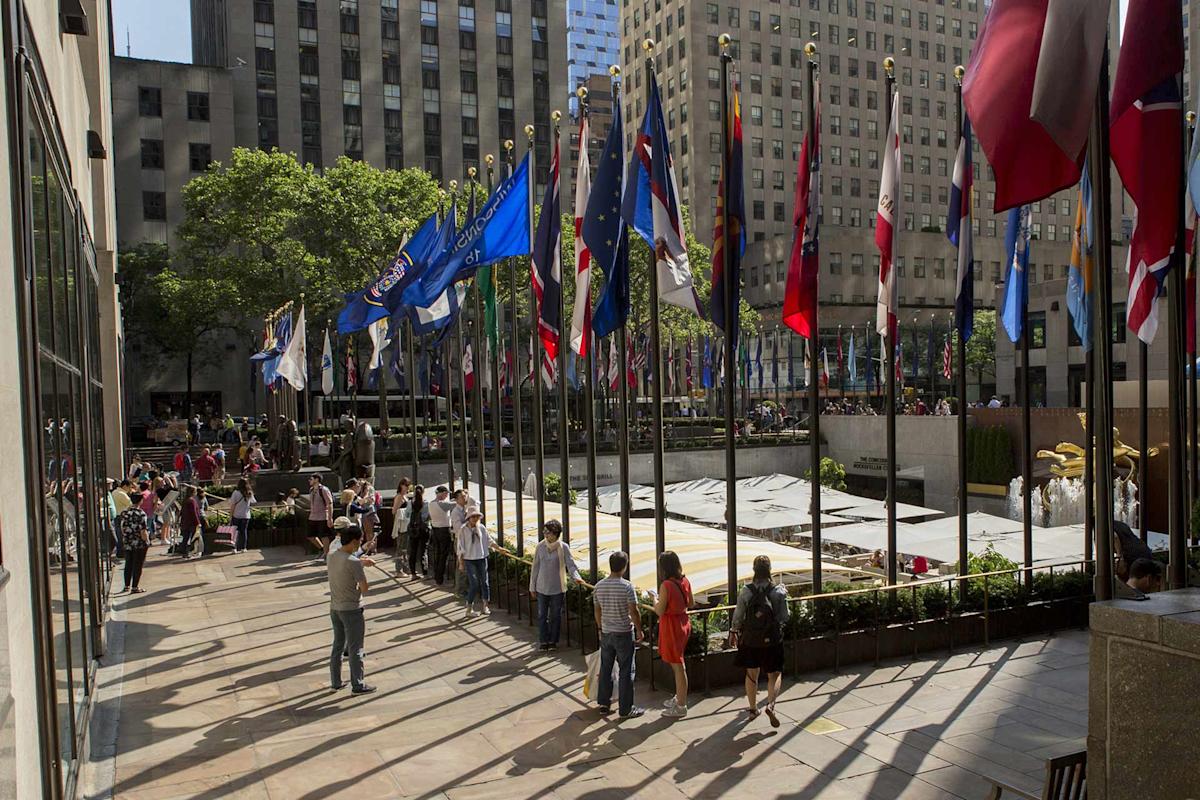 People walk along a plaza lined with many international flags at Rockefeller Center in New York City. Tall buildings and greenery are visible in the background under clear, sunny skies.
