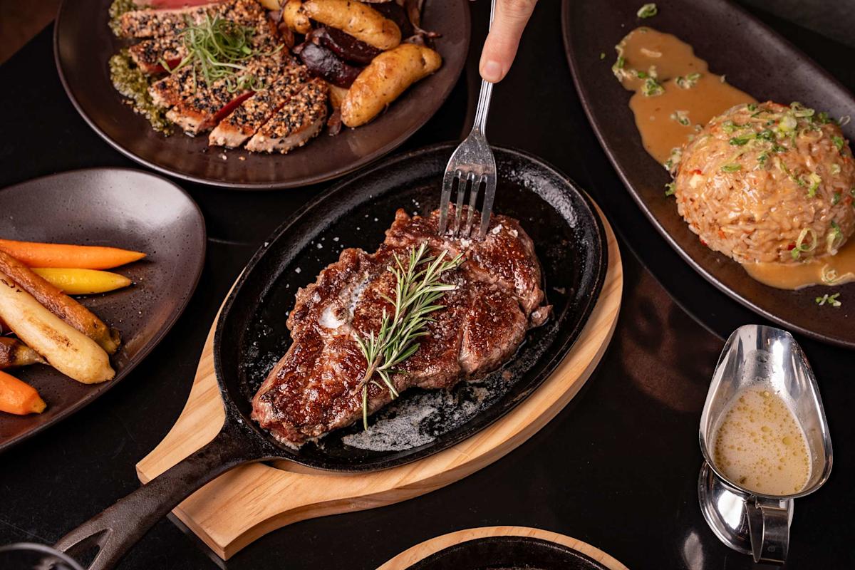 A hand uses a fork to hold a sizzling grilled steak with rosemary on a hot plate, surrounded by dishes of roasted vegetables, rice, and sliced meat with sauce.