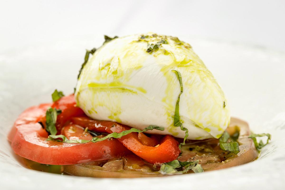 A close-up of a burrata cheese ball topped with olive oil and chopped herbs, served over sliced tomatoes and red peppers on a white plate.