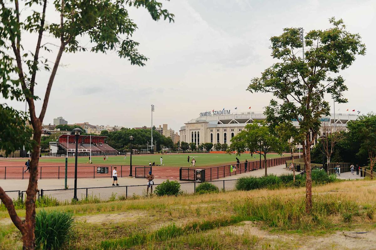 A sports field with people walking and playing, surrounded by trees and greenery, with Yankee Stadium visible in the background under a cloudy sky.