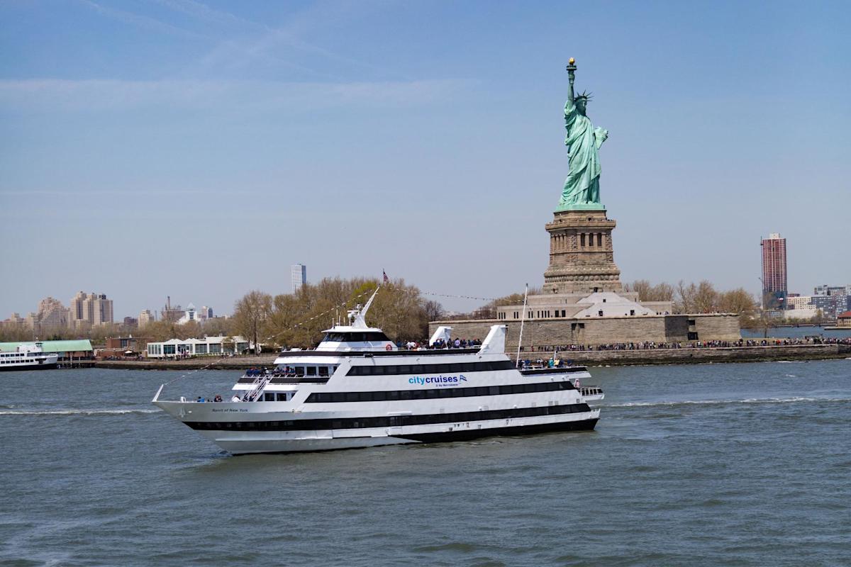 A white City Cruises ferry boat sails past the Statue of Liberty on a sunny day, with passengers on deck and the New York City skyline in the background.