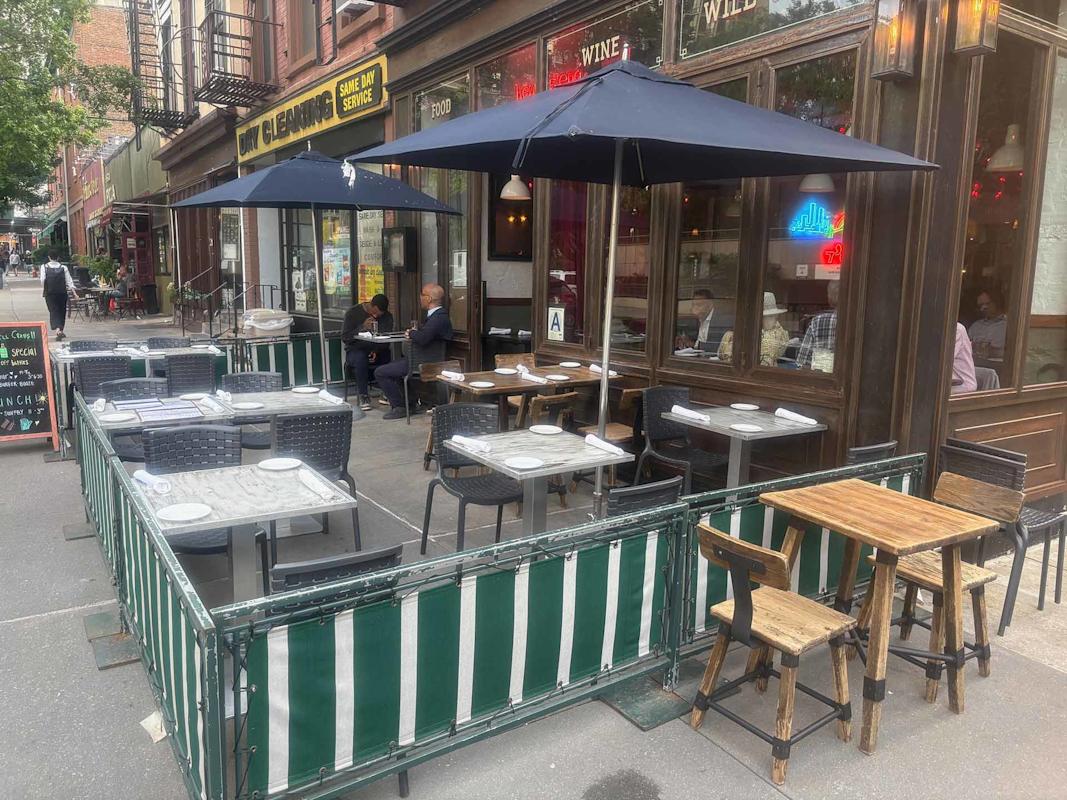 Outdoor seating area of a restaurant with tables and chairs, some set for dining. Two people sit at one table. Green and white barriers enclose the area. Building storefronts and sidewalk visible in the background.