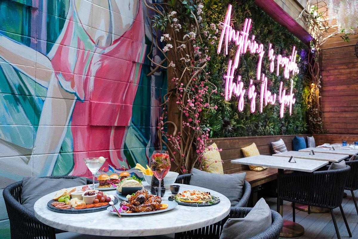 A marble table with assorted dishes and drinks is set in a stylish outdoor café, featuring a mural of a figure and a neon sign reading “Anything is possible, this is New York” on a lush, plant-covered wall.