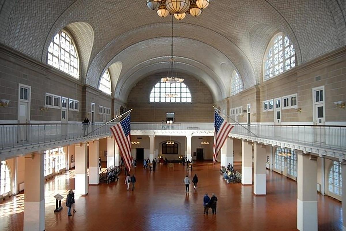 Large, bright hall with arched windows, vaulted ceilings, chandeliers, and two large American flags. People are walking and standing throughout the spacious, sunlit interior.
