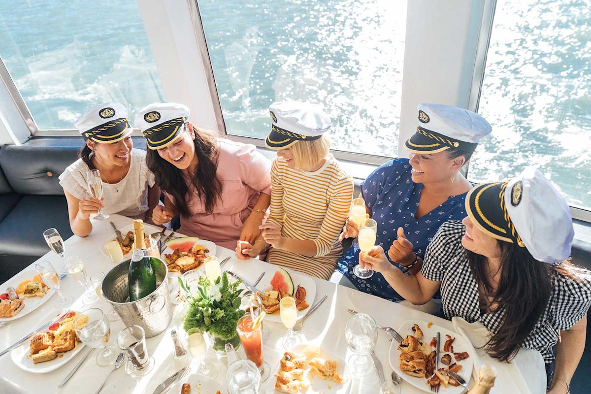 Five women wearing captain hats sit around a table on a boat, laughing and enjoying food and drinks with a view of sparkling water through the windows behind them.