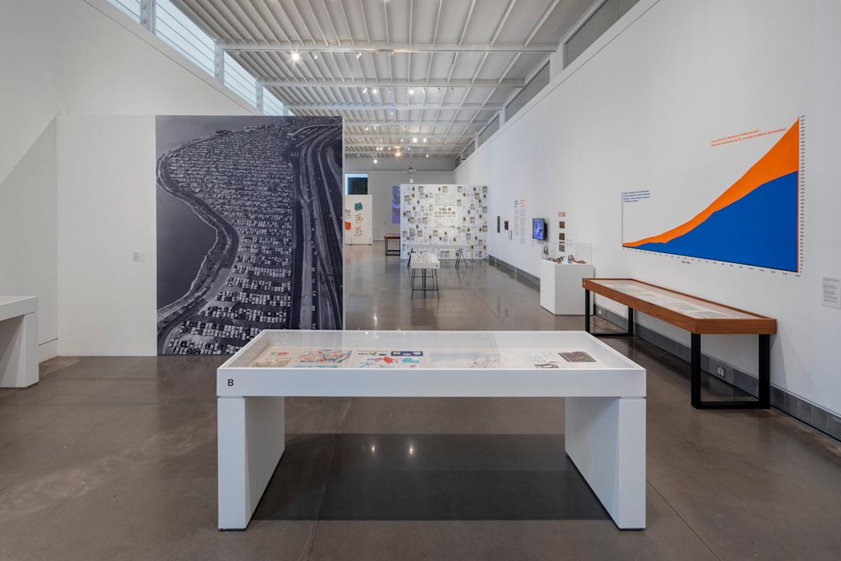 A modern art gallery with a large black-and-white aerial photo on one wall, a colorful line graph on another, and display tables featuring various artworks. The room has high ceilings with skylights and a polished concrete floor.