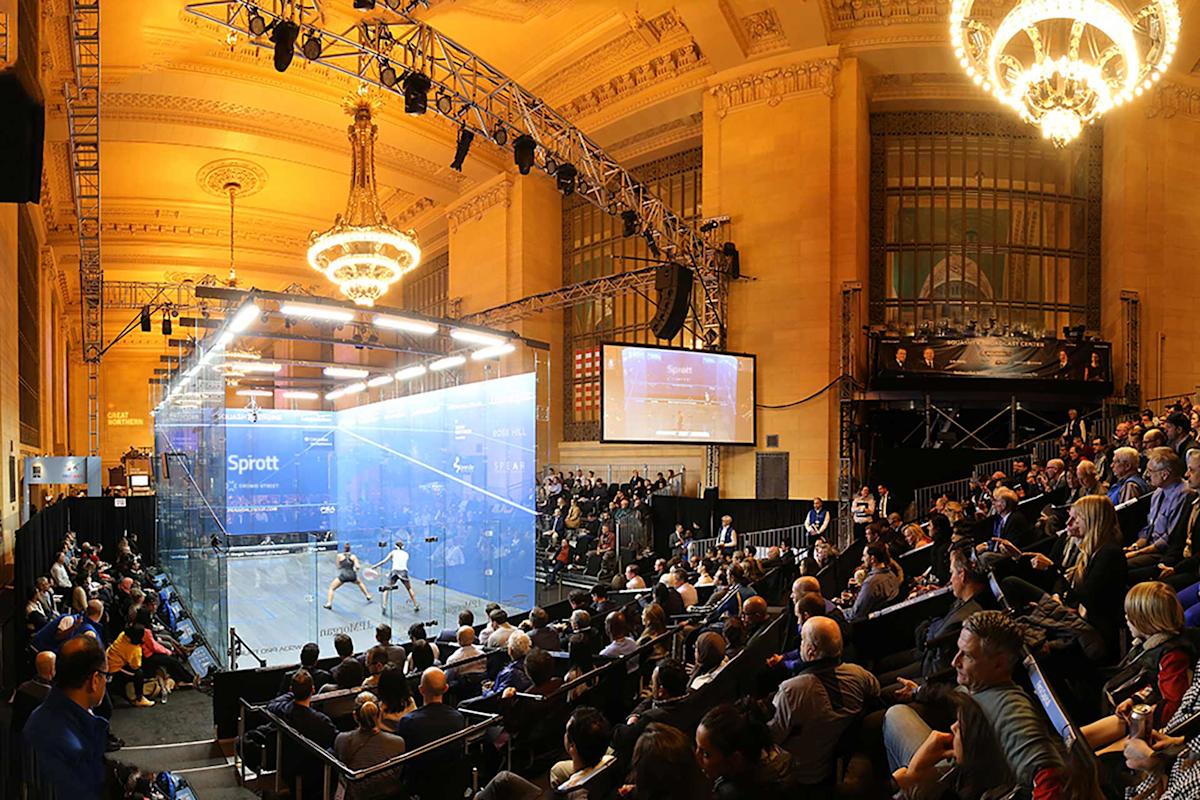 A large audience watches a squash match in a glass court set up inside an ornate hall with chandeliers, high ceilings, and grand windows. A screen displays the game, and lighting rigs hang above the court.