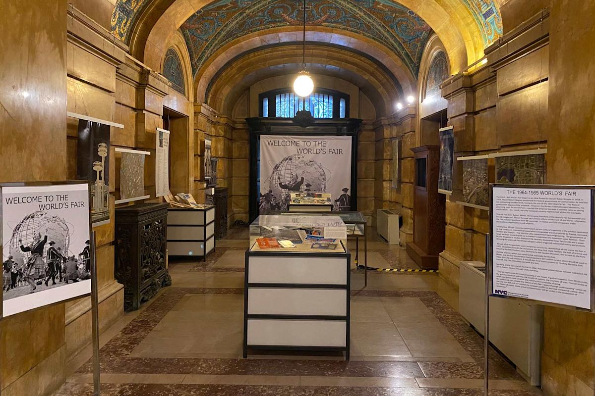A historical exhibit in a grand hall with an arched ceiling. Posters and display cases feature "Welcome to the World's Fair" signage. The walls are ornate, and a single round light hangs from above, creating a warm ambiance.