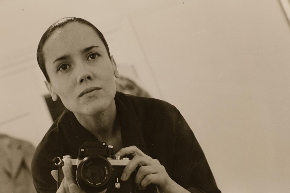 A woman with dark hair pulled back holds a camera and looks into a mirror, capturing her reflection in a sepia-toned photograph. She wears a dark top and has a thoughtful expression.