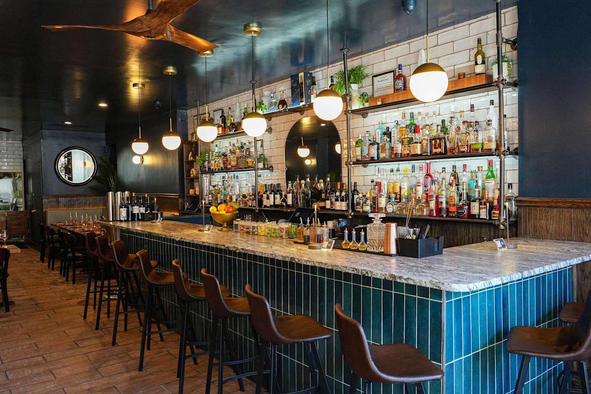 A modern bar with green-tiled counter, brown leather barstools, hanging globe lights, and shelves filled with liquor bottles against a white tiled wall. Wooden tables and chairs line one side of the room.