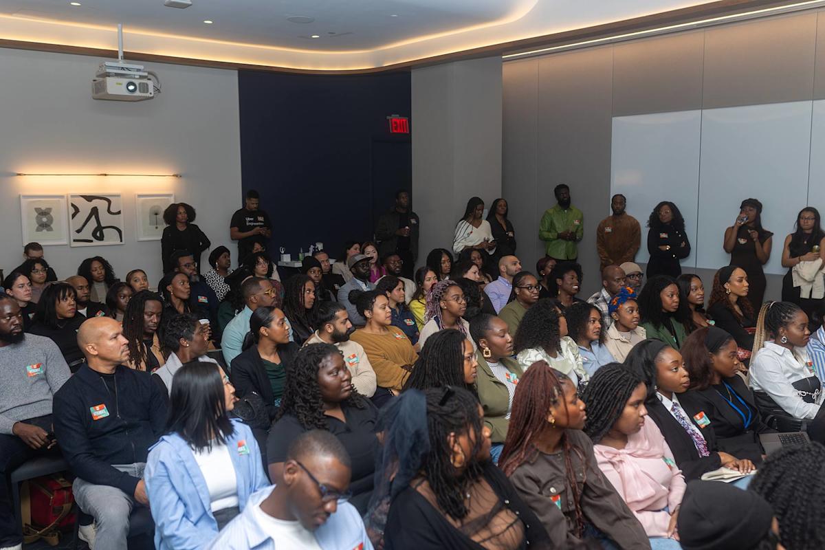 A diverse group of people sit attentively in a modern conference room, facing a speaker out of frame. Some stand along the walls, and a few people have name tags. The atmosphere appears focused and engaged.