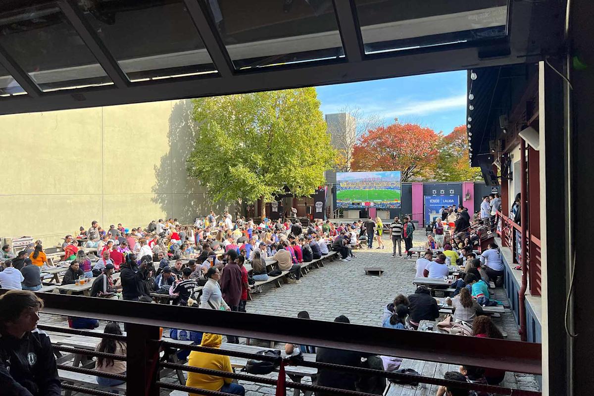 A large crowd sits at outdoor picnic tables watching a sporting event on a big screen in a courtyard, surrounded by trees with green and autumn-colored leaves. Some people are standing or walking by.
