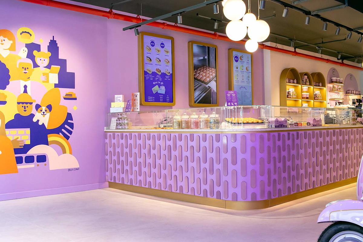 A brightly lit dessert shop with pastel purple and yellow decor, a candy jar-filled counter, colorful wall art, menu boards, and pastries on display under glass domes.