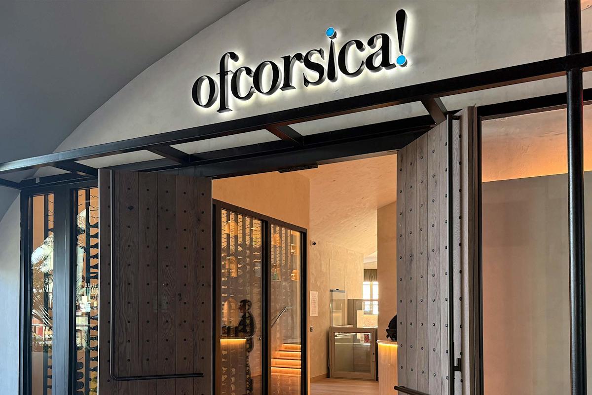 Entrance to a modern restaurant or business with a sign above the door that reads "ofcorsica!" in bold, illuminated letters. The open doors reveal a warm, inviting interior with wood and glass accents.