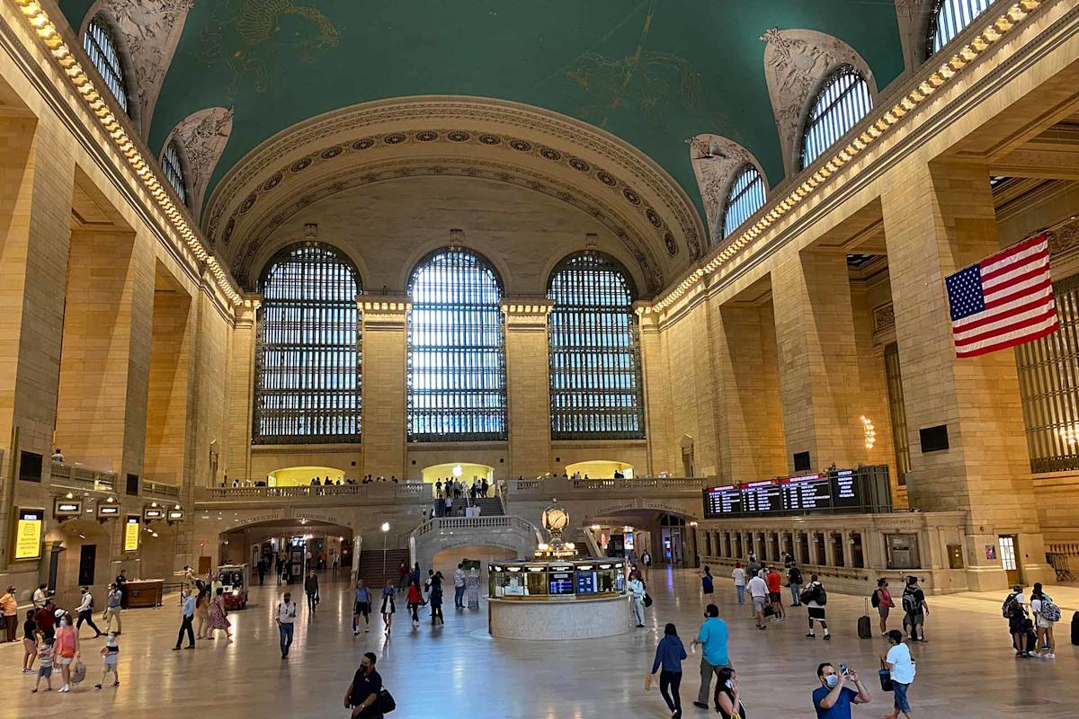 Interior of Grand Central Terminal with high arched windows, a green ceiling with constellations, people walking on the marble floor, an information booth in the center, and an American flag on the right.