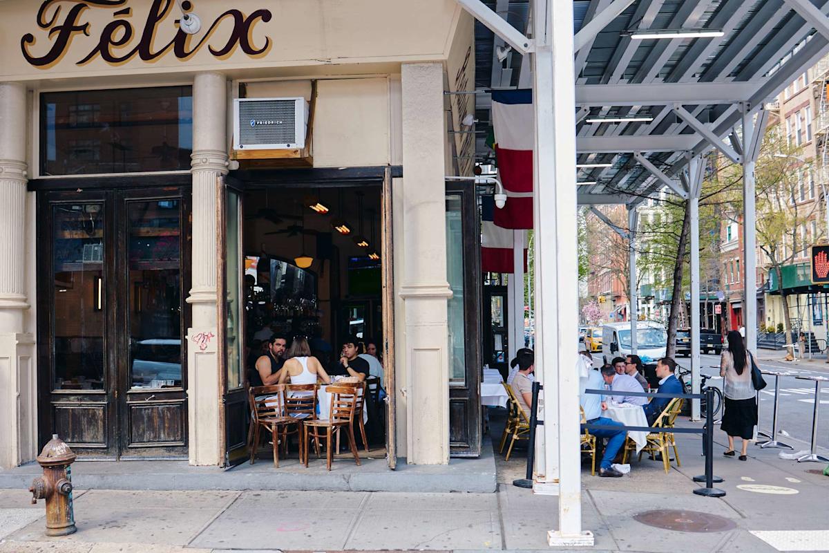 People are dining both inside and outside a corner restaurant called Félix. The street has tables with customers, a sidewalk hydrant, and scaffolding overhead. Trees and city buildings line the background.