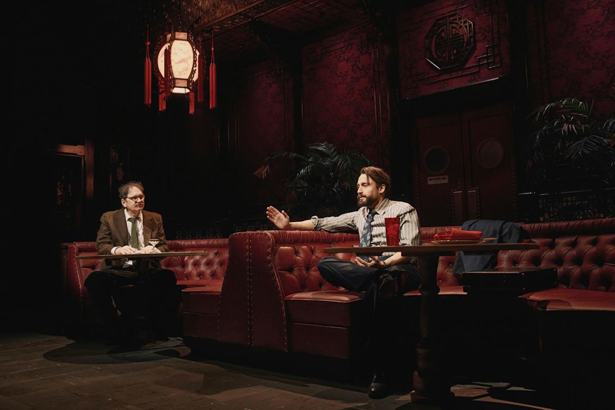Two men sit on red tufted booths in a dimly lit, ornate restaurant or bar. One gestures while speaking; the other listens. The setting features dark decor, a large lantern, and plants in the background.