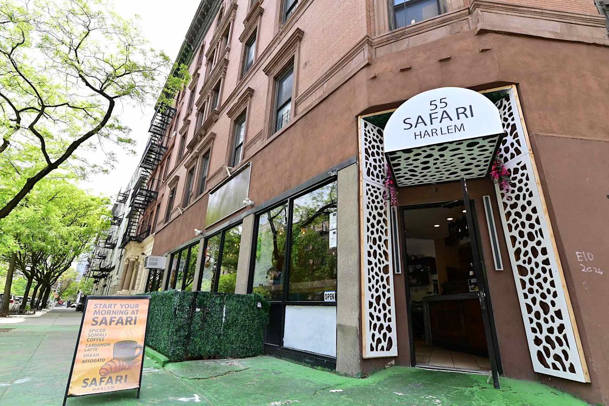 Street view of Safari Harlem at 55, with a white sign above the entrance. An outdoor sign promotes breakfast options. The building is brown with large windows, and trees line the sidewalk.