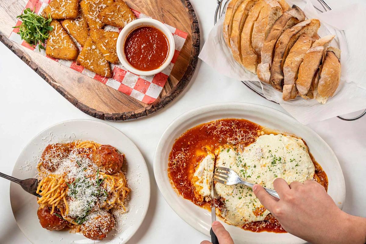 A table with plates of Italian food: bread slices, fried mozzarella sticks with marinara sauce, spaghetti with meatballs, and a person cutting into cheesy baked lasagna in tomato sauce.