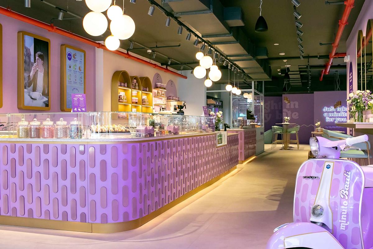 A modern, brightly lit dessert shop with a purple and gold color scheme, curved counters, jars of sweets, decorative lights, and a purple scooter parked inside near pastel seating.