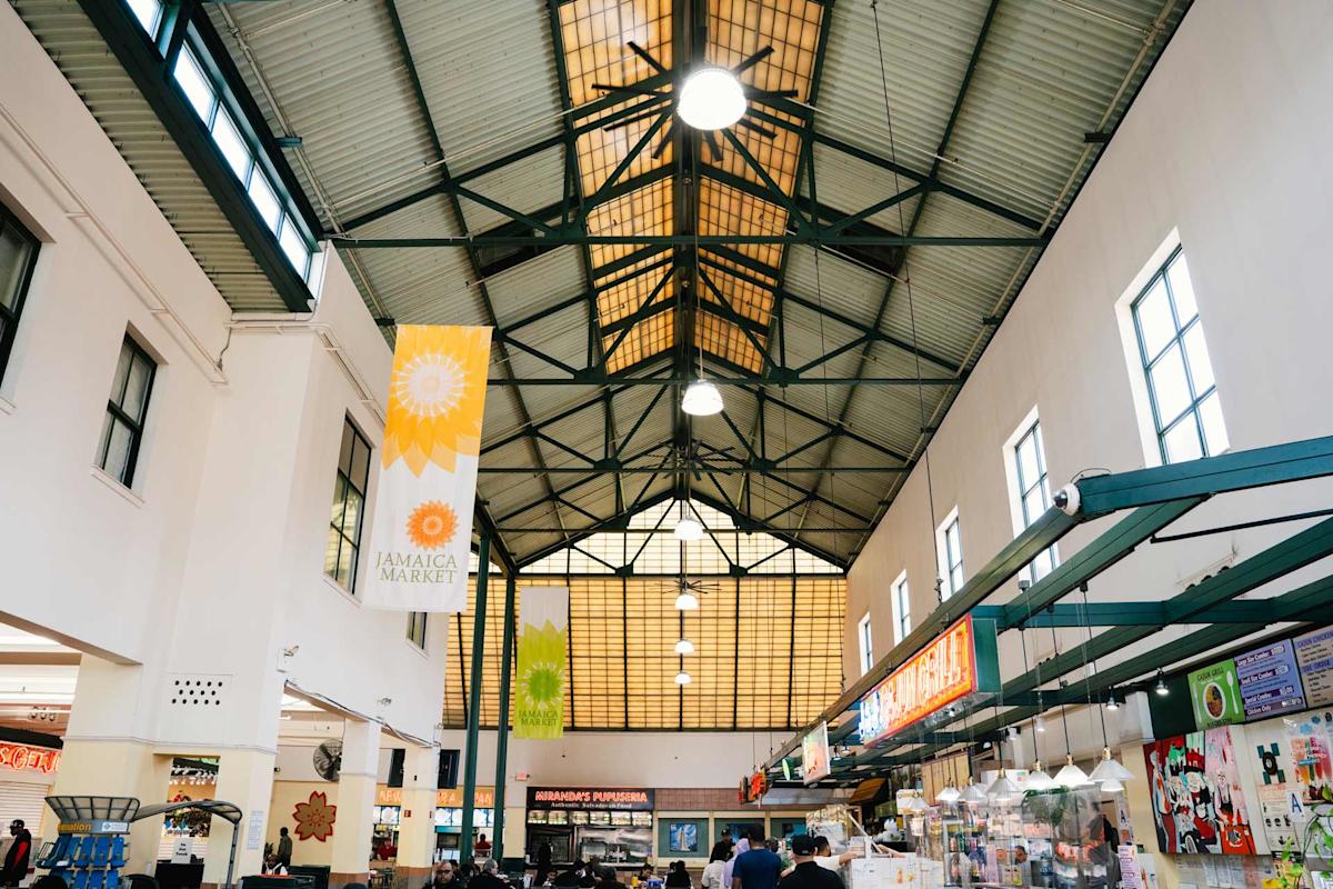 Indoor view of Jamaica Market with a high, triangular ceiling, hanging banners, food stalls on the right, and people walking around. Bright natural light comes through large windows at the end.