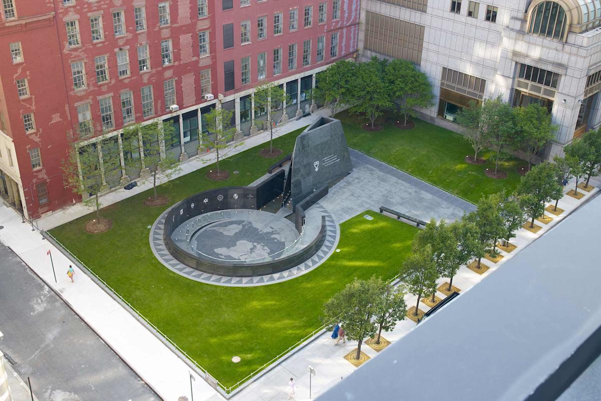 Aerial view of the African Burial Ground National Monument in New York City. The site features a circular memorial with symbolic engravings, surrounded by green lawns and trees, set between red and gray buildings.
