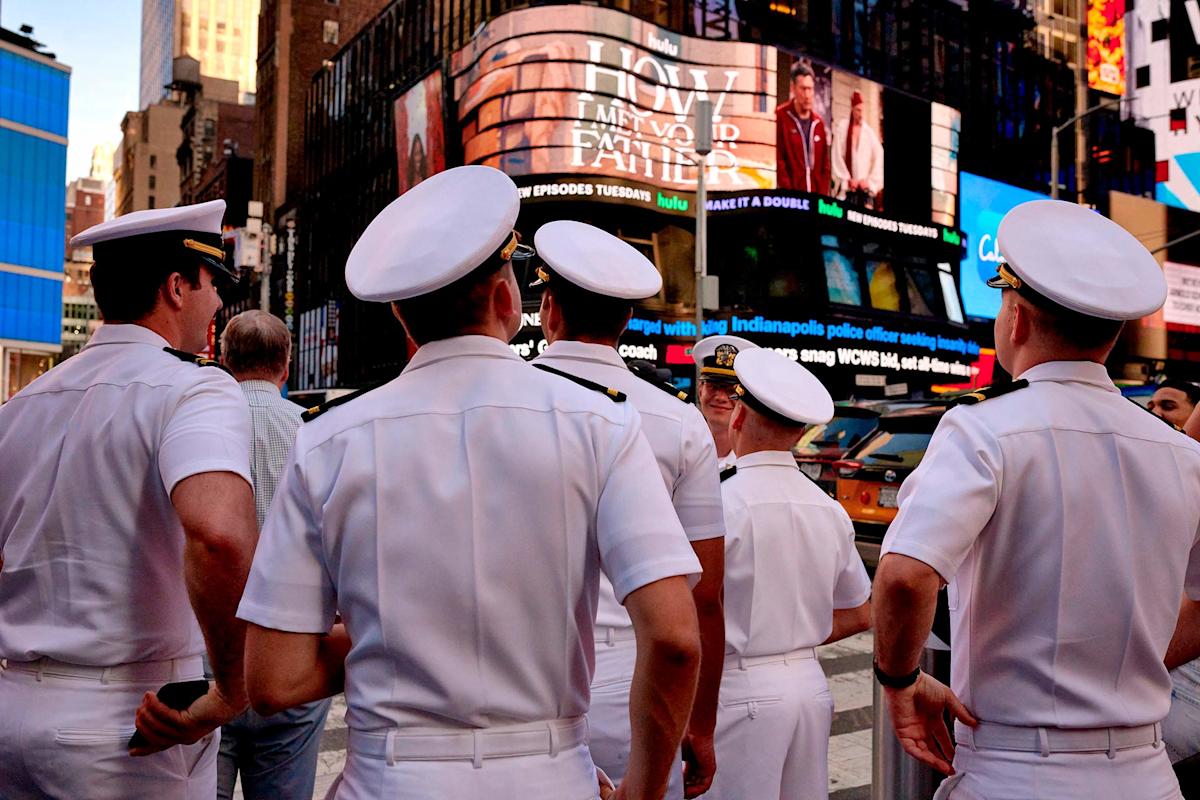 A group of people in white naval uniforms stand together in a busy urban area, with bright electronic billboards and tall buildings in the background.