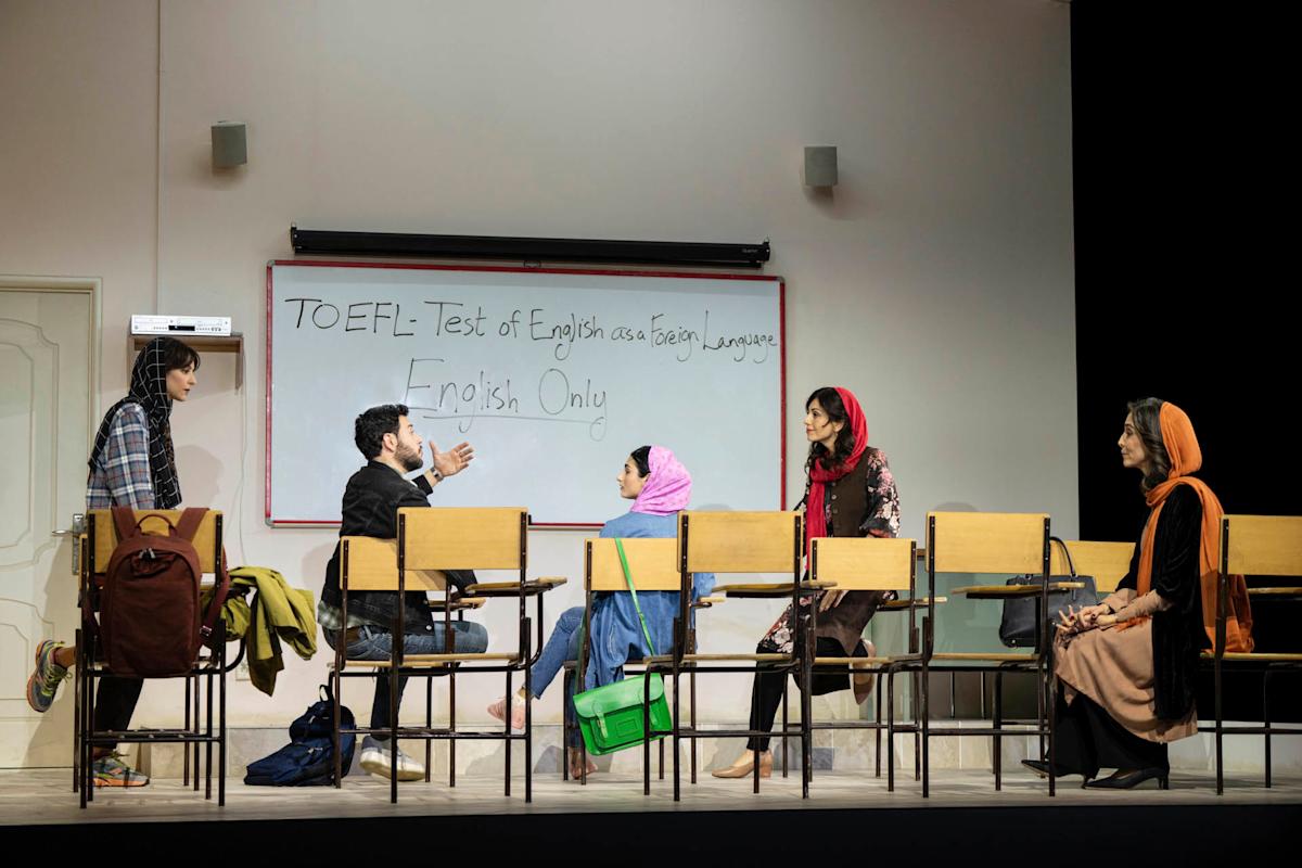 Five people sit and talk in a classroom setting with chairs arranged in a row. A whiteboard behind them reads "TOEFL: Test of English as a Foreign Language. English Only." Some people wear headscarves.