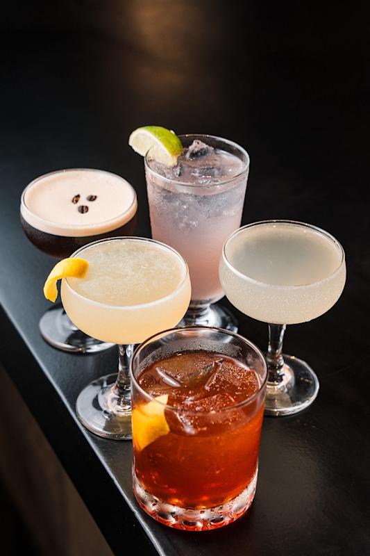 Five assorted cocktails in various glasses sit on a dark surface. Some are garnished with lemon or lime, coffee beans, or served over ice, showcasing a range of colors and styles.