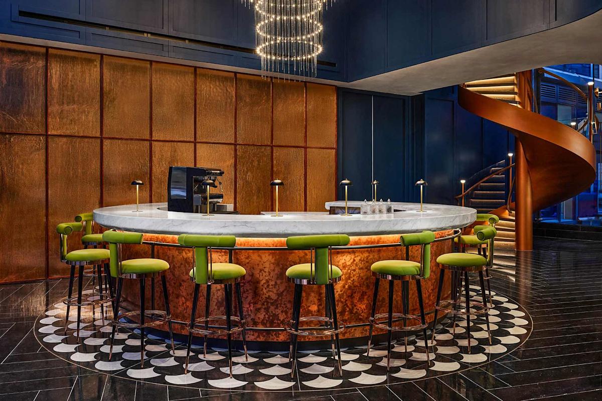 A modern bar with a white marble countertop, green bar stools, copper paneling, black tile floor with a circular pattern, and a spiral staircase in the background under a chandelier.