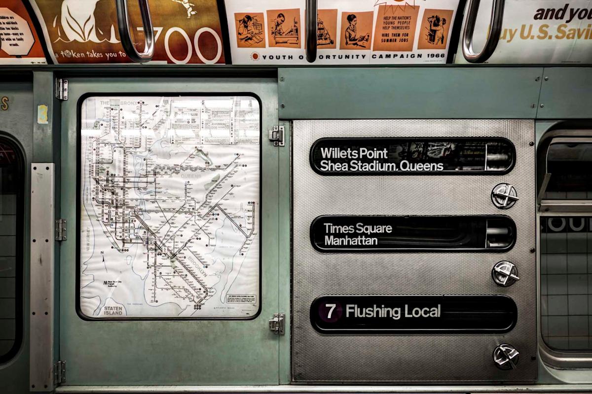 Vintage subway car interior with a black-and-white transit map on the left side and destination signs on the right for Willets Point, Times Square, and Flushing Local. The setting has a retro design with beige overhead advertisements.